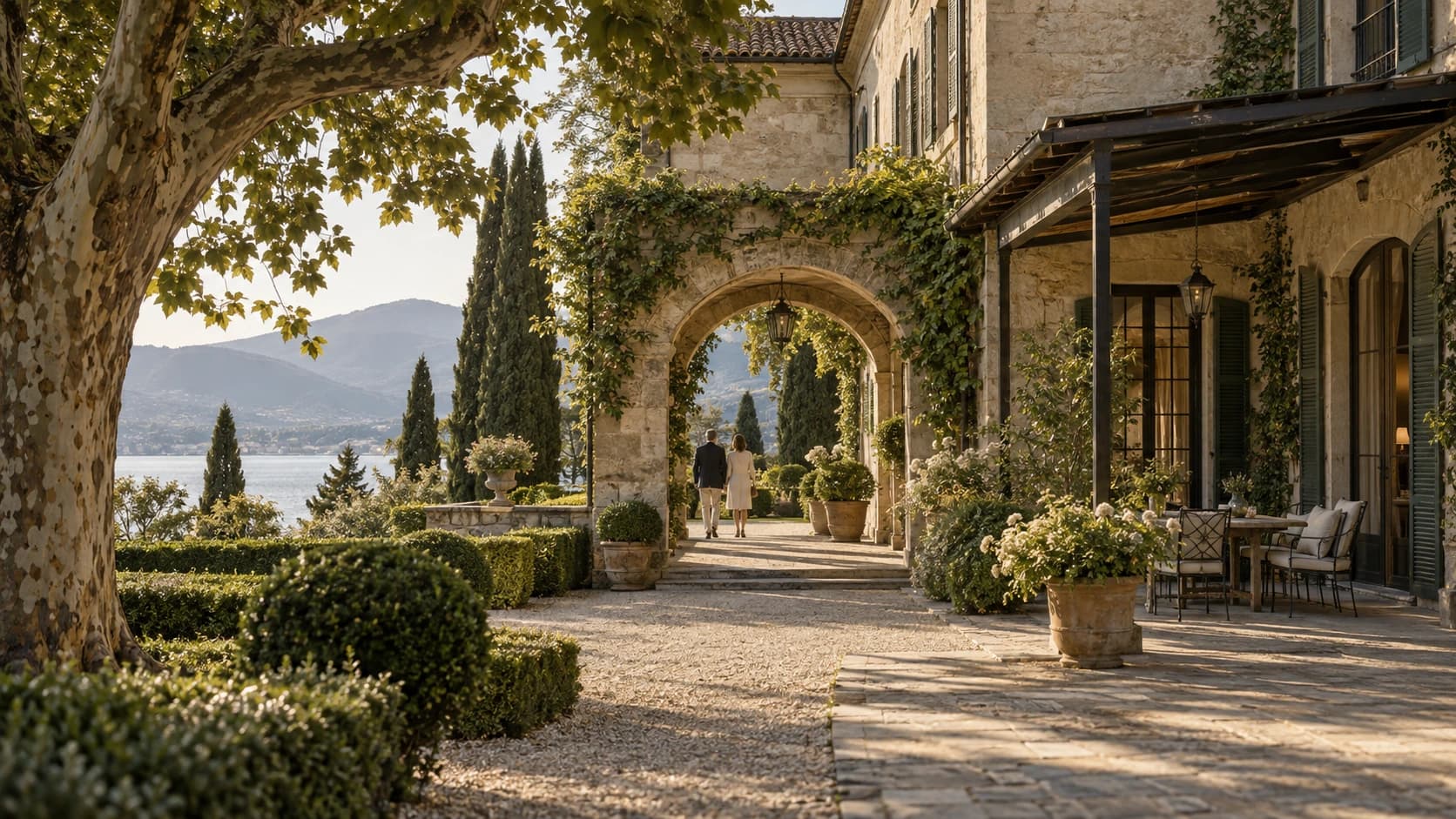 A sunlit limestone villa beside a lake, with mature gardens and two people walking beneath a stone arch.